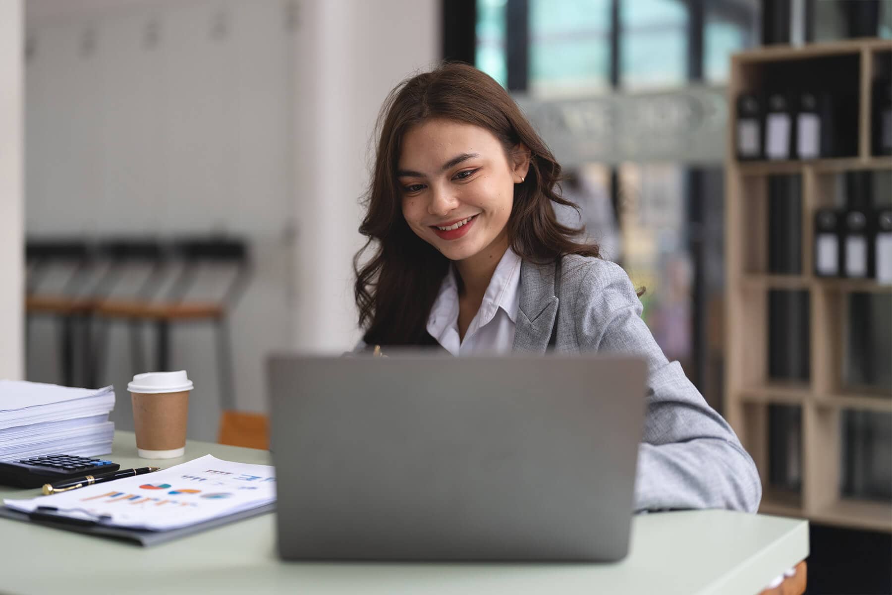 Smiling woman working on laptop with documents and coffee in modern office space.